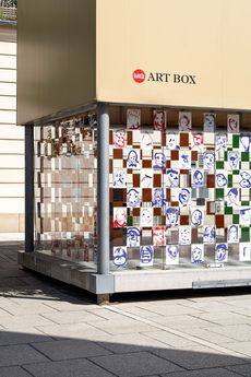 Kunstinstallation im öffentlichen Raum mit dem Namen „MQ ART BOX“. Zu sehen ist eine gläserne Fassade, die mit einem Raster aus verschiedenen Porträtzeichnungen und einem Schachbrettmuster verziert ist und vor einem modernen urbanen Hintergrund steht.