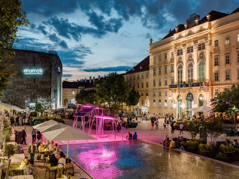 Blick auf den belebten Innenhof des MQ mit Gastronomie und bunter Beleuchtung bei Dämmerung.