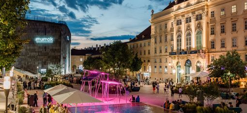 View of the bustling courtyard of the MQ with restaurants and colorful lighting at dusk.