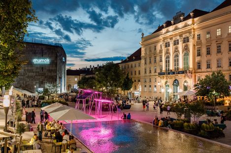 Blick auf den belebten Innenhof des MQ mit Gastronomie und bunter Beleuchtung bei Dämmerung.