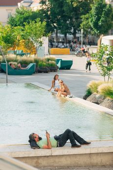 Menschen entspannen an einem flachen Wasserbecken in einem begrünten Stadtplatz. Ein Mann liegt am Rand und schaut auf sein Smartphone, während zwei Frauen mit den Füßen im Wasser sitzen. Im Hintergrund sind Bäume, Sitzgelegenheiten – die MQ Enzis – und weitere Besucher zu sehen.