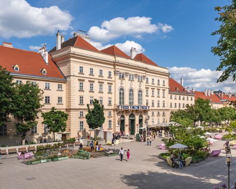 Large historic building with red roofs and several trees in front on a busy square with seating and people