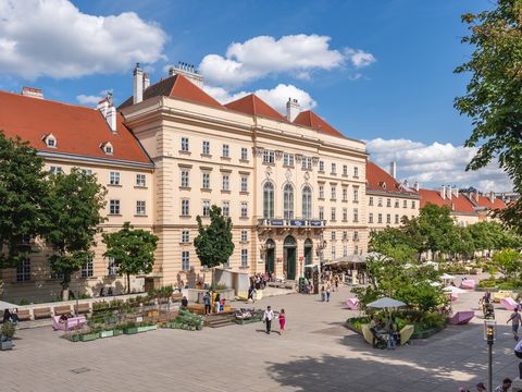 Large historic building with red roofs and several trees in front on a busy square with seating and people