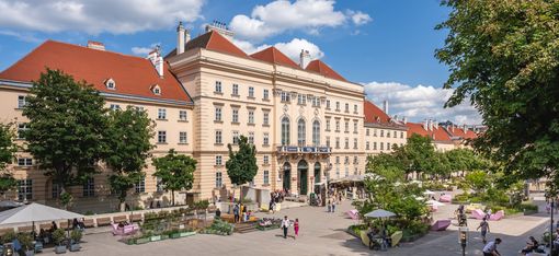 Large historic building with red roofs and several trees in front on a busy square with seating and people