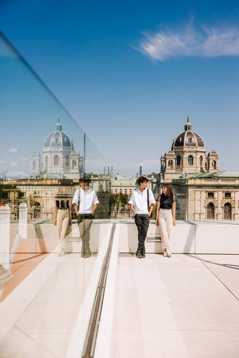 Ein junges Paar steht auf einer Dachterrasse und blickt auf die Skyline einer Stadt, wobei ein großes Gebäude mit Kuppel im Hintergrund dominiert und sich in einer Glasfläche spiegelt.
