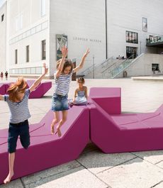 Drei Kinder in gestreiften Oberteilen spielen auf modularen, pinkfarbenen Sitzmöbeln vor dem Leopold Museum.