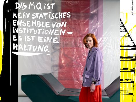 Frau mit rotem Rock und lila Jacke steht vor einer modernen, geometrischen Sitzbank vor einer roten und weißen Wand mit dem Text 'DAS MQ IST KEIN STATISCHES ENSEMBLE VON INSTITUTIONEN ES IST EINE HALTUNG'