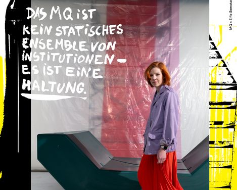 Woman wearing a red skirt and purple jacket standing by a modern geometric bench in front of a red and white wall with the text 'DAS MQ IST KEIN STATISCHES ENSEMBLE VON INSTITUTIONEN ES IST EINE HALTUNG'