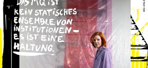 Frau mit rotem Rock und lila Jacke steht vor einer modernen, geometrischen Sitzbank vor einer roten und weißen Wand mit dem Text 'DAS MQ IST KEIN STATISCHES ENSEMBLE VON INSTITUTIONEN ES IST EINE HALTUNG'