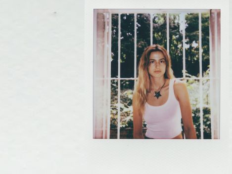 Young woman with long hair wearing a white tank top sitting in front of a window with bars