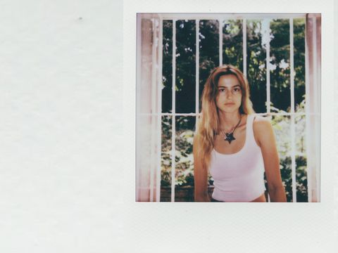 Young woman with long hair wearing a white tank top sitting in front of a window with bars