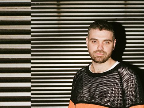 Man with short hair and beard wearing a mesh shirt with an orange stripe standing in front of a wall with horizontal stripes