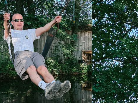 Mann mit Sonnenbrille schaukelt an einer Kette vor grünem Laub und einem Holzgebäude