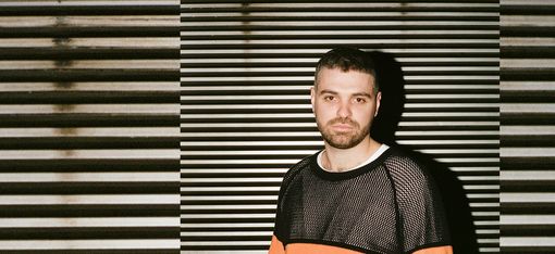 Man with short hair and beard wearing a mesh shirt with an orange stripe standing in front of a wall with horizontal stripes