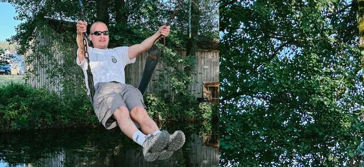 Man wearing sunglasses swinging on a chain in front of green foliage and a wooden building