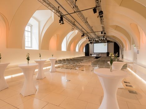 Veranstaltungsraum Arena21 im MuseumsQuartier mit gewölbter Decke, Reihen von Stühlen vor einer Leinwand und Stehtischen mit weißen Hussen und einzelnen Blumenvasen.