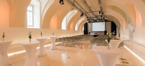 Veranstaltungsraum Arena21 im MuseumsQuartier mit gewölbter Decke, Reihen von Stühlen vor einer Leinwand und Stehtischen mit weißen Hussen und einzelnen Blumenvasen.