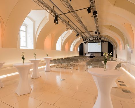 Veranstaltungsraum Arena21 im MuseumsQuartier mit gewölbter Decke, Reihen von Stühlen vor einer Leinwand und Stehtischen mit weißen Hussen und einzelnen Blumenvasen.