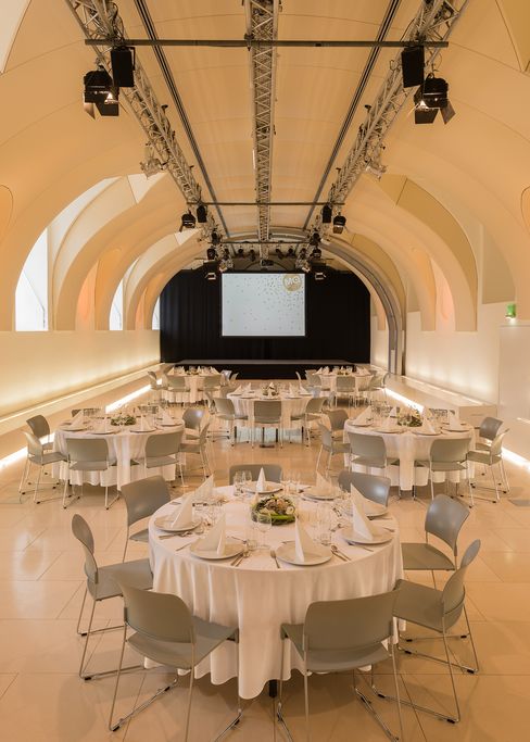 Großer Veranstaltungsraum Arena21 im MuseumsQuartier Wien mit gewölbter Decke, runden Tischen mit weißen Tischdecken und gedeckten Plätzen, Stühlen und einer Leinwand am Ende des Raums