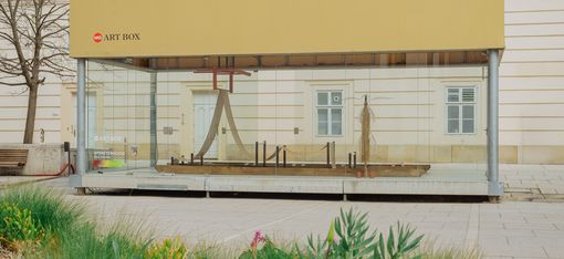 Exterior view of the MQ Art Box in MuseumsQuartier Vienna, showing the installation inside with greenery in the foreground.
