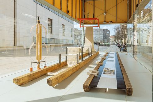 Interior view of the exhibition featuring long wooden beams, perforated metal surfaces, and a sculptural construction inside a glass pavilion.