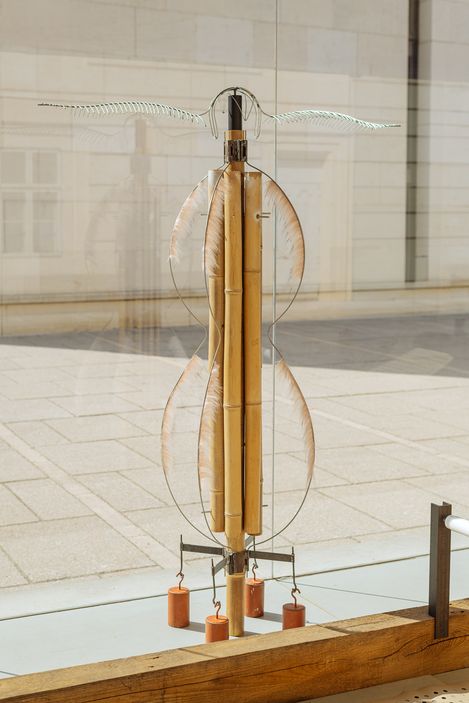 Close-up of a delicate sculpture made of bamboo, feathers, and wire, displayed behind glass in the MQ Art Box pavilion.