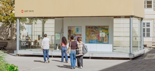 Four people walking toward a modern glass building with artworks displayed in the window