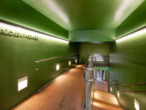 Interior view of a green bridge passage with stairs and ramp, illuminated by wall lights and featuring the word »BRÜCKENPASSAGE« on the wall