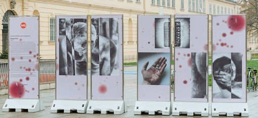 Freestanding multi-panel photographic installation in MuseumsQuartier Vienna featuring fragmented black-and-white images of the human body with red spots on a light background.