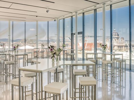 Innenraum der Eventlocation MQ Libelle im MuseumsQuartier mit hohen Tischen und Hockern vor bodentiefen Fenstern, die einen Blick über Wien bieten.