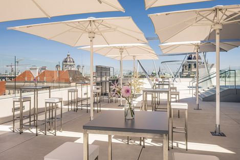 MQ Libelle Dachterrasse mit Tischen, Stühlen und großen Sonnenschirmen, im Hintergrund Blick auf Wiener Gebäude und blauen Himmel.