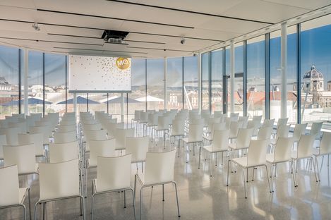 Moderner Veranstaltungsraum mit weißen Stühlen vor einer Leinwand, große Fenster bieten Blick auf die Dächer und Gebäude Wiens.