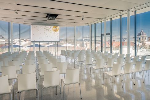Moderner Veranstaltungsraum mit weißen Stühlen vor einer Leinwand, große Fenster bieten Blick auf die Dächer und Gebäude Wiens.