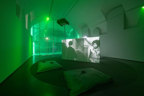 Dark exhibition room with green lighting, a large screen showing a black-and-white video, and two cushions on the floor