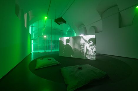 Dark exhibition room with green lighting, a large screen showing a black-and-white video, and two cushions on the floor