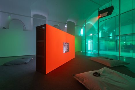 Large orange room divider with a screen in a dim exhibition space with cushions on the floor and glass walls in the background