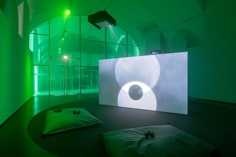 Dark exhibition room with green lighting, a large screen showing an abstract image, and two floor cushions with headphones in front