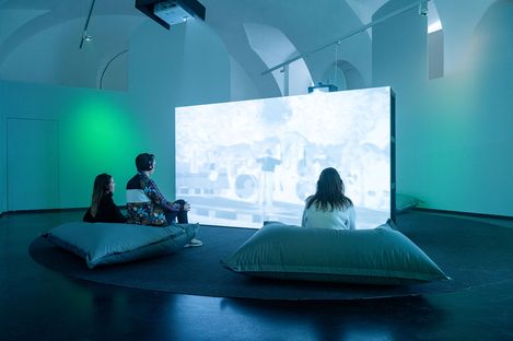 Three people sit on large cushions facing a bright screen in a dark room with vaulted ceiling