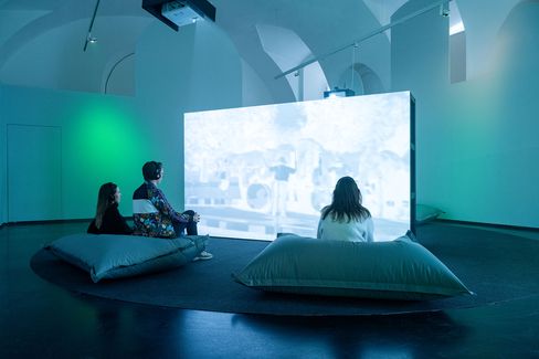 Three people sit on large cushions facing a bright screen in a dark room with vaulted ceiling