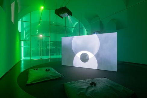 Dark exhibition room with green lighting, a large screen showing an abstract image, and two floor cushions with headphones in front