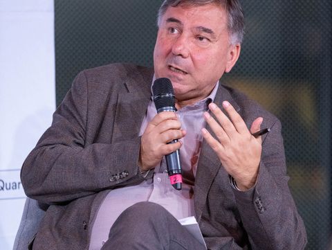 Mann sitzt mit verschränkten Beinen auf einem Sessel und hält ein Mikrofon in der rechten Hand und einen Stift in der linken Hand