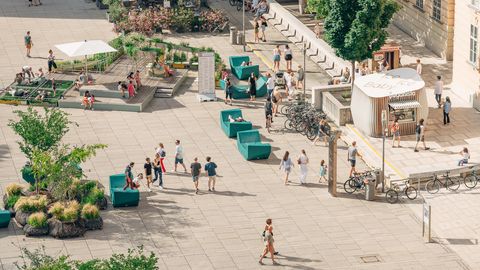 Blick von oben auf einen belebten Platz mit Sitzbänken, Fahrradständern, Pflanzen und Menschen, die sich bewegen oder sitzen