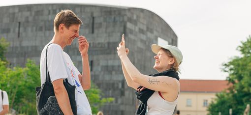 Zwei Menschen lachen miteinander im MQ Haupthof, während eine Person die andere mit dem Smartphone fotografiert. Im Hintergrund sind das mumok, Besucher:innen und die grünen Enzis des MuseumsQuartier Wien zu sehen.