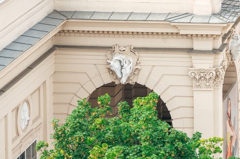 Architekturdetail eines historischen Gebäudes mit Säulen, Ornamenten und einem Wappen über einem Bogen, davor ein grüner Baum