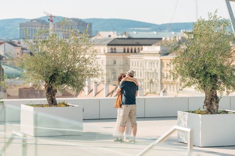 Paar umarmt sich auf der Dachterrasse der MQ Libelle mit großen weißen Pflanzkübeln und Stadtblick im Hintergrund.
