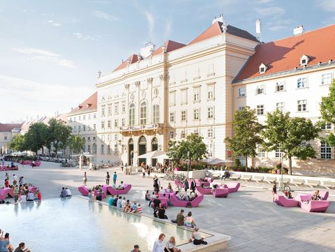 Menschen sitzen und verweilen rund um das Wasserbecken im Haupthof des MuseumsQuartier Wien, umgeben von pinken Enzi-Sitzmöbeln und der historischen Architektur der ehemaligen Hofstallungen bei sommerlichem Wetter.