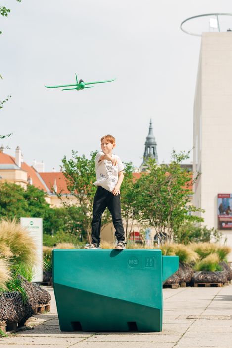 Child standing on a green Enzi throwing a toy airplane, with the MuseumsQuartier in the background
