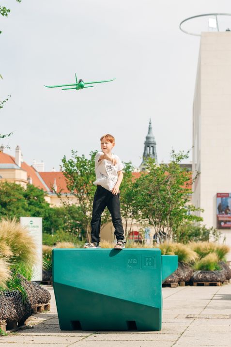 Child standing on a green Enzi throwing a toy airplane, with the MuseumsQuartier in the background