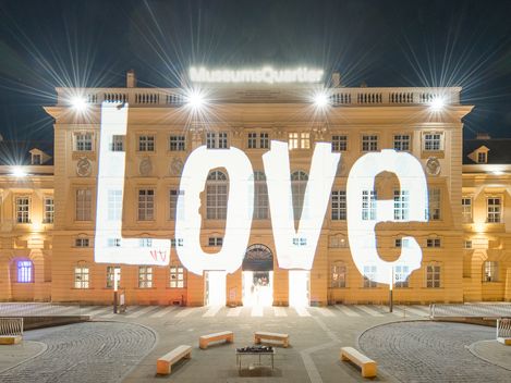 The facade of a historic building, the MQ forecourt, at night. Bright light projections illuminate the words “everything for everyone” and “everything” in various sizes on the facade.