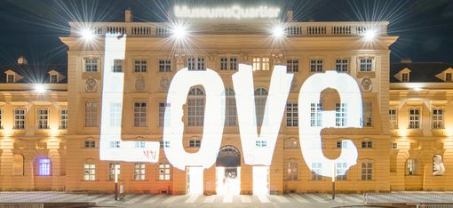 The facade of a historic building, the MQ forecourt, at night. Bright light projections illuminate the words “everything for everyone” and “everything” in various sizes on the facade.